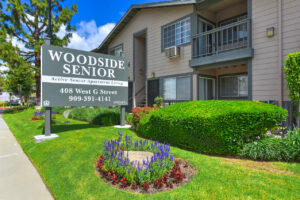 Exterior Woodside Senior Front Signage, meticulously groomed grounds, flowers planted along sidewalk, photo taken on a sunny day with clouds.