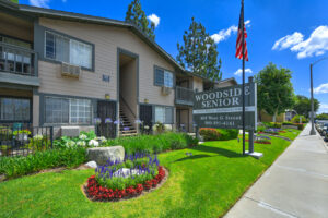 Exterior Woodside Senior, Entrance Signage, Flowers planted along sidewalk, meticulous landscaping, beige building with dark green trim, photo taken on a sunny day.