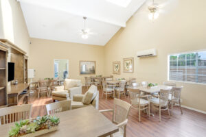 Interior Clubhouse, beige walls with wood floors, large rustic tv console with bookshelves, multiple tables with 6 seats at each table along wall, large beige lounge chairs.