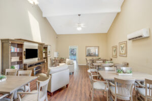 Interior Clubhouse, beige walls with wood floors, large rustic tv console with bookshelves, multiple tables with 6 seats at each table along wall, large beige lounge chairs.