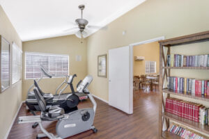 Interior Fitness room, treadmill, elliptical machine, stationary bike, bookshelf in corner of the room, wood floors, beige walls, windows with shutters, door to community room.