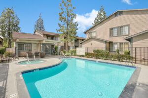 Exterior Woodside Pool, pool area with hot tub, chairs around pool area, pool area fenced in, beige buildings with dark green trim in background, photo taken on a sunny day.