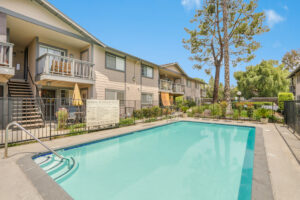 Exterior Woodside Pool, Beige building with dark green trim in background, lounge chairs along pool, lush foliage, photo taken on a sunny day.