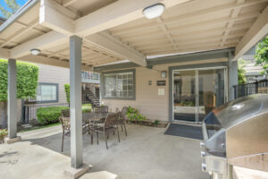 Exterior Clubhouse, covered round table with 5 seats, gas grill, beige building with dark green trim, meticulous landscaping in the background.