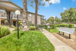 Exterior Woodside residential building, Beige building with dark green trim, Meticulous landscaping, bench along sidewalk, lush foliage, photo taken on a sunny day.